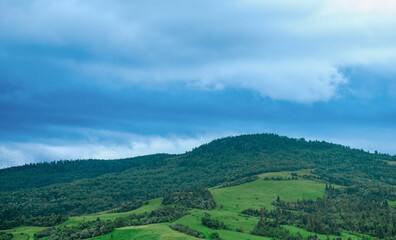Fototapeta premium High-quality landscape of green rolling hills and mountains under a dramatic blue cloudy sky in summer.