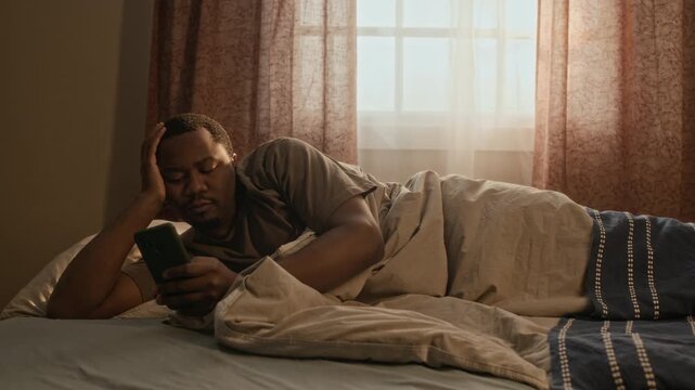 Medium long shot of young African American man lying in bed and scrolling on smartphone with tired expression. Video is ideal for mental health awareness, sleep disorder or digital detox campaigns