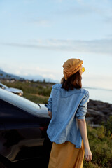 Fototapeta premium Young woman in a denim shirt and hat stands beside a parked car by the coast, capturing a casual outdoor lifestyle moment with soft daylight and distant sea.