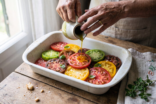 Mains d'une femme &acirc;g&eacute;e versant de l'huile d'olive sur des tranches de tomates rouges, vertes et jaunes, feuilles de basilic et de sauge