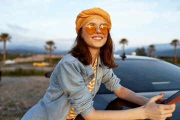 Fototapeta premium A smiling woman wearing an orange headscarf and denim jacket leans on a car in a sunny outdoor setting, showcasing casual lifestyle, travel, and carefree moment.