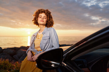 Fototapeta premium A woman with curly hair wearing a denim jacket and striped top stands beside a car at sunset, showcasing a relaxed, outdoor lifestyle moment suitable for travel and leisure themes.