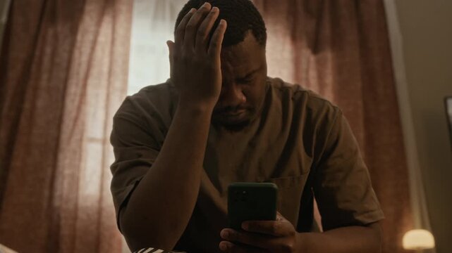 Zoom in shot of young African American man sitting on bed, holding head in distress while looking at smartphone. Clip is perfect for mental health campaigns or digital fatigue concepts