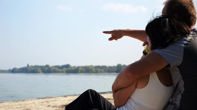 Happy pair hugs during sitting on wooden log on beach. Romantic couple admiring sea landscape at sunny day. Lovers spending time together at resort. Concept of summer vacation or holiday. Slow mo