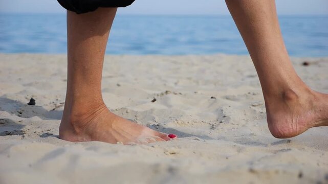 Female feet walking on sand at the beach with seascape at background. Legs of adult woman stepping along sea shore. Barefoot lady strolling near ocean coast. Concept of summer vacation. Low view