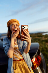 Fototapeta premium Cheerful woman outdoors taking a selfie, smiling with eyes opened, striped shirt, denim jacket, casual fashion, warm sunlight, vibrant colors, natural landscape, car in background, candid lifestyle