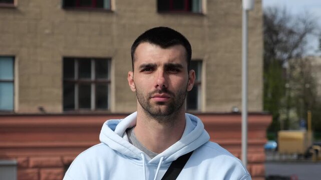 Portrait of young confident bearded man in casual clothing with cityscape background. Close up of handsome guy looks into camera standing at city environment at sunny day
