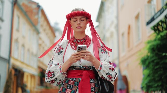 Portrait of a woman dressed in a traditional Ukrainian vyshyvanka, red floral wreath, and beaded necklaces, focused on her smartphone while standing on a historic cobblestone city street.