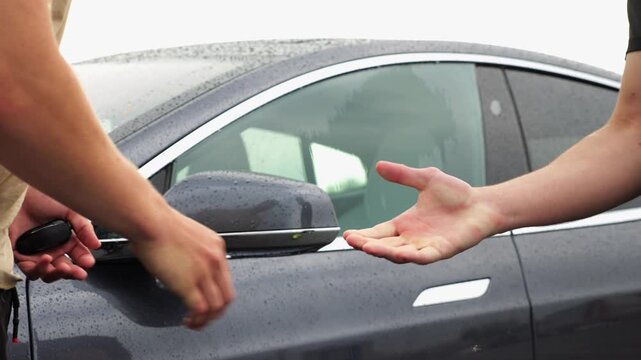 Male arm giving key of car to buyer of electric automobile at rainy day. Handshake between salesman and new owner of modern auto after successful deal. Concept of buy or rent car. Slow motion