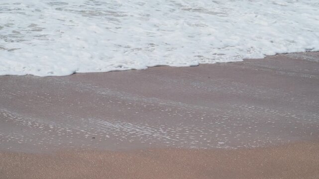 Close up of calm ocean waves roll toward sandy shore. White foam meets warm sand at shoreline. Gentle water motion creates natural rhythm. Perfect for serene vacation or relaxation scenes