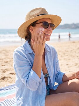 Beautiful young woman in a straw hat sitting on a sandy beach and applying sunscreen lotion to her cheek on a sunny summer day