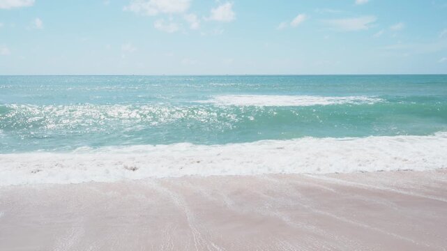 Clear blue sky with fluffy white clouds. Bright turquoise ocean stretches to the horizon. Gentle waves for white, frothy patterns. Ripples move slowly across the sandy shore