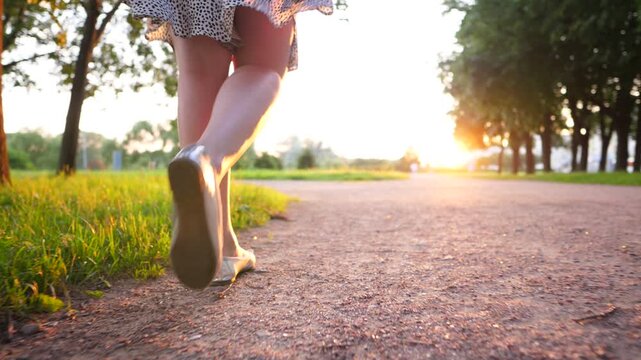 Slender legs of young woman, walking along gravel path in city park, slow motion follow camera. Beautiful evening sun light, bright shine ahead. Grass on left and some trees beside alley