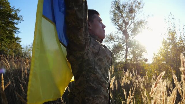 Military man in uniform stands with raised over head flag of Ukraine at countryside. Male ukrainian army soldier with lifted national banner in honor of victory against russian aggression. End of war