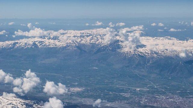 Stunning mountain landscape from airplane window. High altitude aerial view of white clouds over snow capped peaks and vast blue horizon landscape.