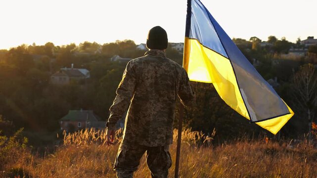 Soldier of ukrainian army going with lifting blue-yellow banner at sunset. Military man in uniform kissing blue-yellow flag and raising it up at countryside. Victory against russian aggression concept