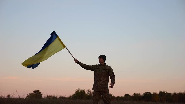 Male military waving blue-yellow banner on liberated territory occupied by russia. Young soldier of ukrainian army lifted flag of Ukraine in honor of victory against russian aggression at dusk