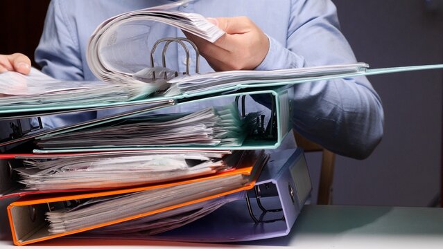 Person arranging and organizing documents within several full binders stacked on a desk, representing a high volume of administrative office work and bureaucracy