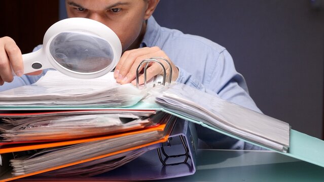 Man holding a magnifying glass, intently reviewing financial documents and binders, symbolizing detailed investigation, deep analysis, and thorough research of important information