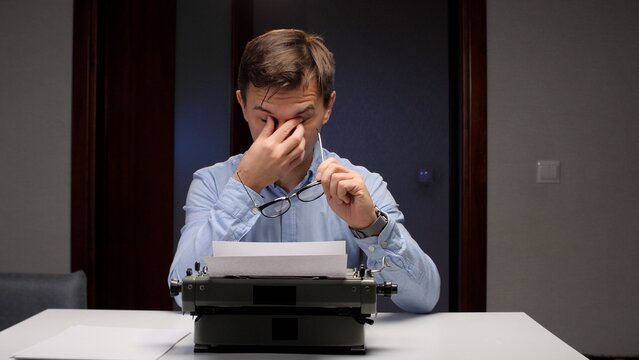 Overworked man rubbing his eyes, holding eyeglasses, and feeling exhausted while trying to type on a blank paper in a vintage typewriter, representing stress and creative fatigue