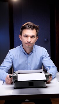Man wearing a blue shirt sitting at a desk, looking at the viewer, with his hands resting on an old fashioned typewriter with a blank sheet of paper, ready for creative writing