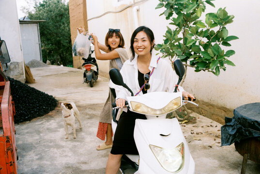 Two Women on Scooters Smiling on a Rural Street