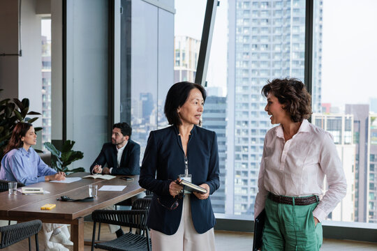 Businesswomen converse in modern office, meeting in background.
