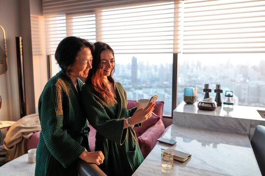 Mother and daughter in robes smile at phone in apartment.