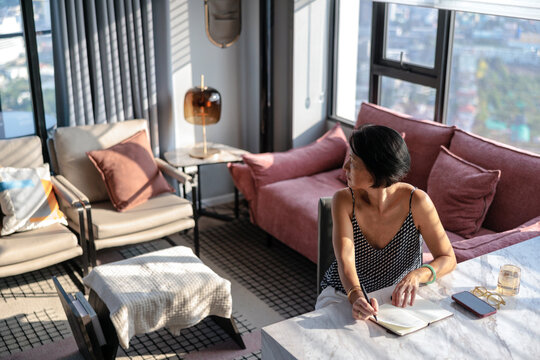 Woman holding pen at table looking over shoulder in sunlit apartment
