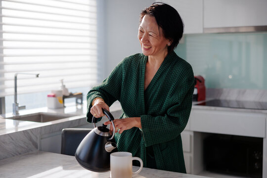 Woman in robe smiles, pouring tea in modern kitchen.
