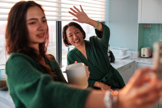 Daughter taking selfie while mother waves in kitchen at home