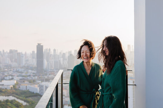 Mother and daughter laugh on balcony in matching robes with city view