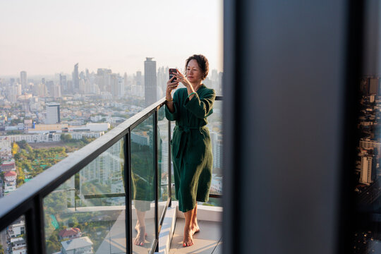 Woman photographing city skyline from balcony in morning robe