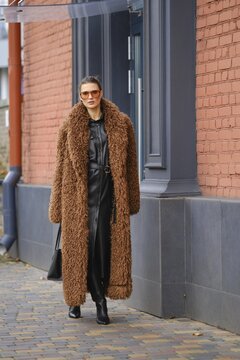A fashionable woman strolls along a city sidewalk, wearing a cozy brown fur coat. She pairs it with a chic black outfit, sunglasses, and confidently walks past a striking red brick building