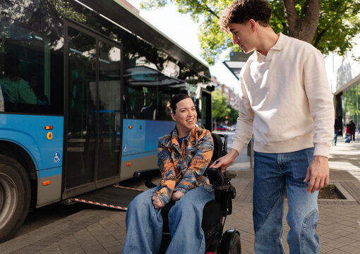 Young man helping woman in electric wheelchair boarding public bus
