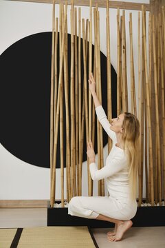A woman bends down to touch bamboo sticks arranged in a room with a large black circle on the wall. The setting conveys an indoor space with natural elements and simple decor