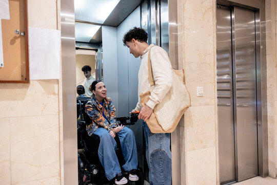 Young man assisting woman in electric wheelchair getting in elevator