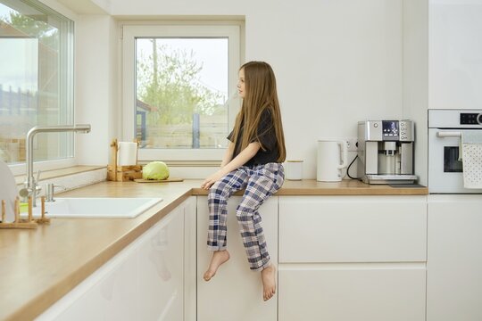 Dreamy young girl in plaid pants and black shirt sits on kitchen countertop and watching to the window