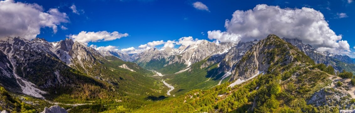 Wide-screen high-resolution panoramic view of the Albanian Alps along the Theth to Valbona hiking route and the Peak of the Balkans region. The landscape features dramatic mountain peaks, deep valleys