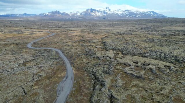 Survol des champs de lave en Islande, travers&eacute; par une route et des montagnes enneig&eacute;es en fond 