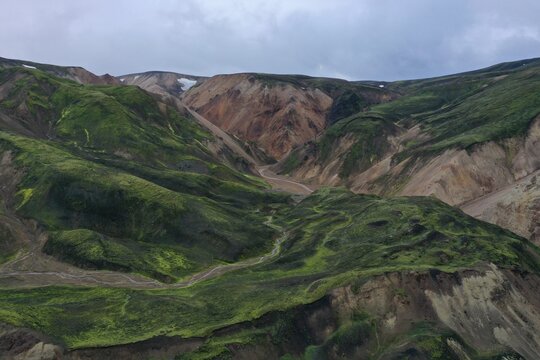 Aerial view of the vibrant green mossy slopes and reddish-brown volcanic terrain of the Kerlingarfjoll mountain range with a winding river in Hella, Rangarthing ytra, Iceland.
