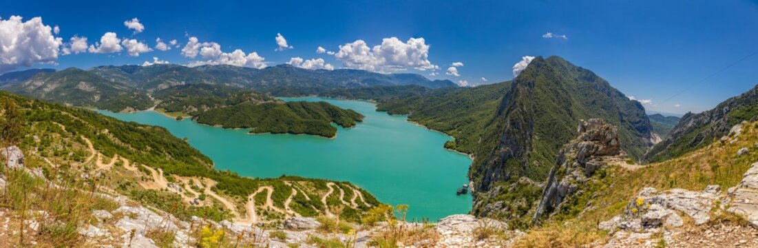 Wide-screen high-resolution panoramic view of Bovilla Lake in Albania, showcasing the striking turquoise waters surrounded by steep rocky cliffs and rugged mountain terrain. The reservoir  Bovilla