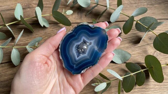 Hand holding a vibrant blue agate geode slice, surrounded by fresh eucalyptus branches, on rustic wooden surface