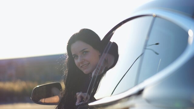 Young beautiful girl leaning out of driver window at modern electric car and looking into camera. Portrait of attractive smiling woman in alternative electrical vehicle. Slow motion