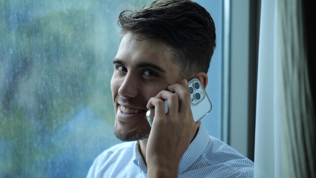 Portrait of male smiling entrepreneur in shirt speaking by smartphone near big window into skyscraper. Happy businessman talking on phone at office. Young manager having conversation on cellphone