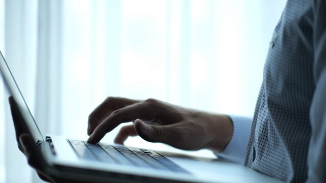 Male hand of young businessman typing text on laptop keyboard near window in modern office. Professional manager working on computer with natural light, showing productivity and communication