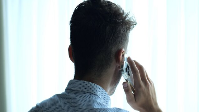 Male entrepreneur in shirt speaking by smartphone near large window into skyscraper. Young businessman talking on phone at office. Confident manager having conversation on cellphone indoor. Close up
