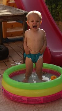 Vertical video of a upset toddler boy crying in a small inflatable swimming pool outdoors. Unhappy little kid throwing a tantrum and pulling his shorts