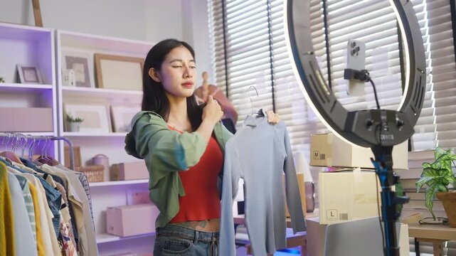 Asian woman live streaming fashion clothing sales with smartphone and ring light, modern retail e-commerce entrepreneur, social commerce influencer and generation z digital side hustle concept