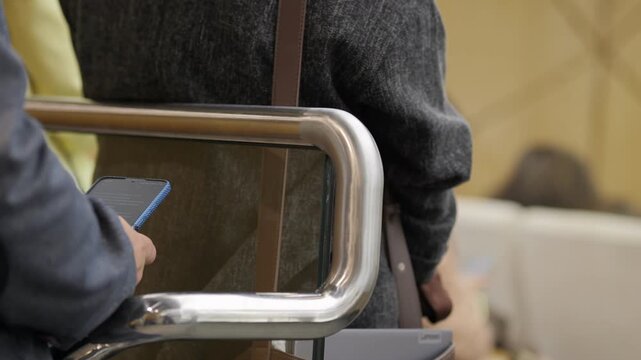 Person holds phone, standing in line at airport gate. Metal railing separates travelers from security zone. Neutral expression on individual's face. Scene captures everyday commute, airport transit